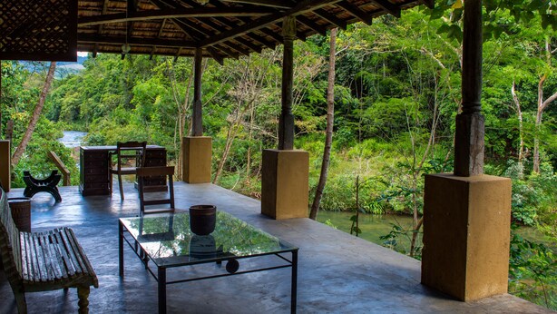 An outdoor seating area with a table and chairs, surrounded by lush greenery and a view of a river. - The Kandy Samadhi Centre (Kundasale)