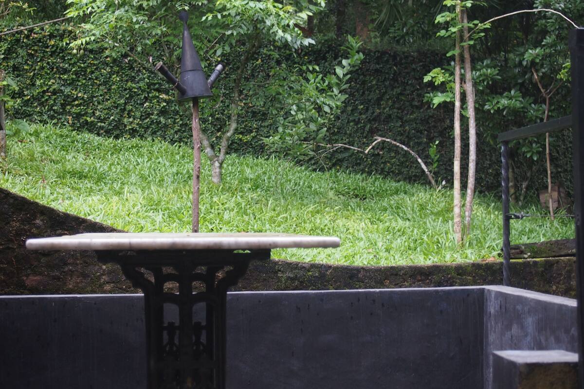 A table with a metal top and legs, surrounded by a low stone wall, set in a garden with grass and trees.