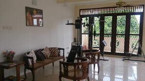 Un salon avec un canapé, des fauteuils et un tapis de course. Une fenêtre offre une vue sur la verdure à l’extérieur.