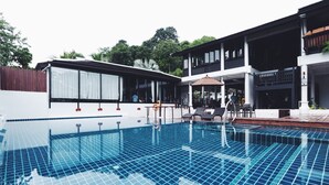 Una piscina con bordeado de baldosas, rodeada por un edificio con varios balcones y un área de estar cubierta.