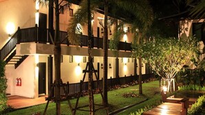 Un edificio de varios pisos con fachada bien iluminada, rodeado de vegetación exuberante y un camino de madera que conduce a la entrada.