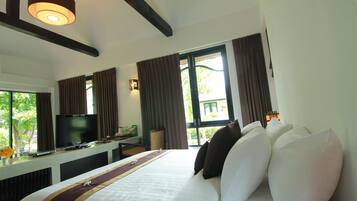A hotel room with a bed, a television, a desk with a computer, a window with curtains, and a view of greenery outside.