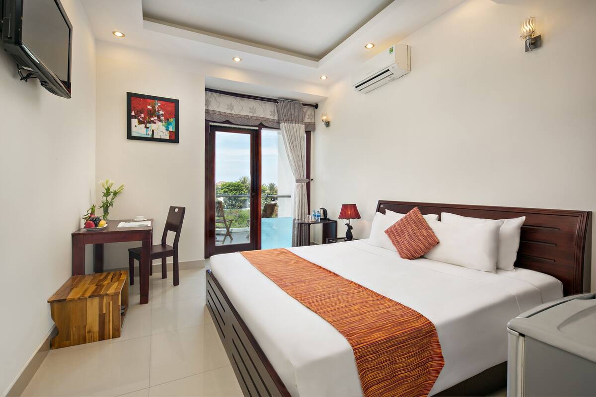 A hotel room with a bed, a desk with a chair, a small table with a plant, and a view through a glass door.
