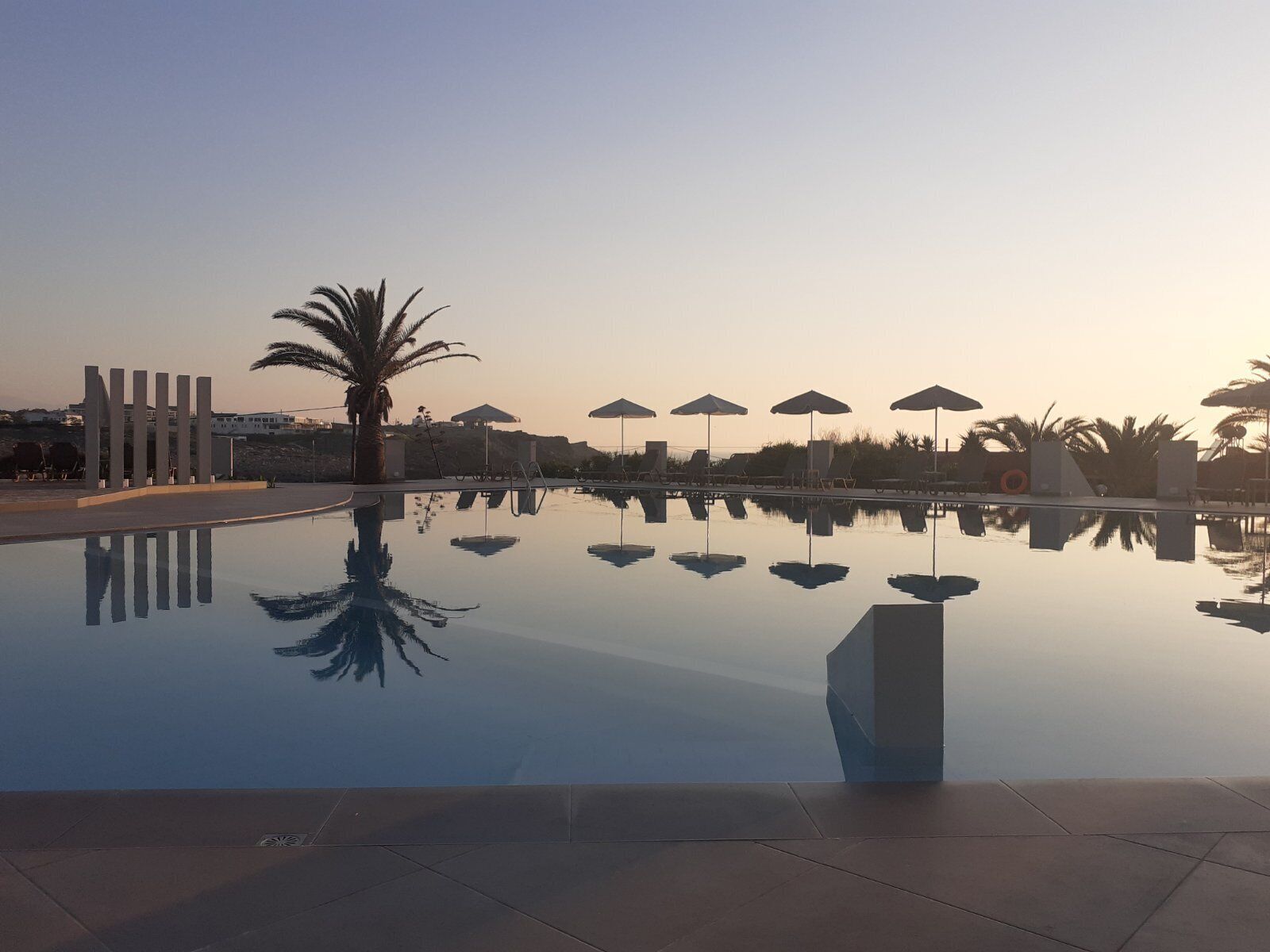 A tranquil pool area with clear reflections, lounge chairs, umbrellas, and palm trees.