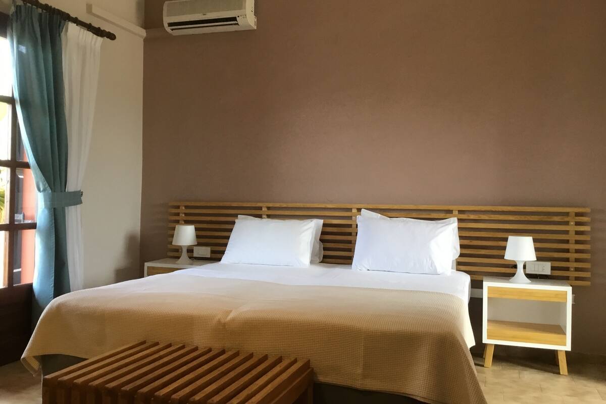 A hotel room with a large bed, two bedside tables with lamps, an air conditioning unit, and a wooden bench.