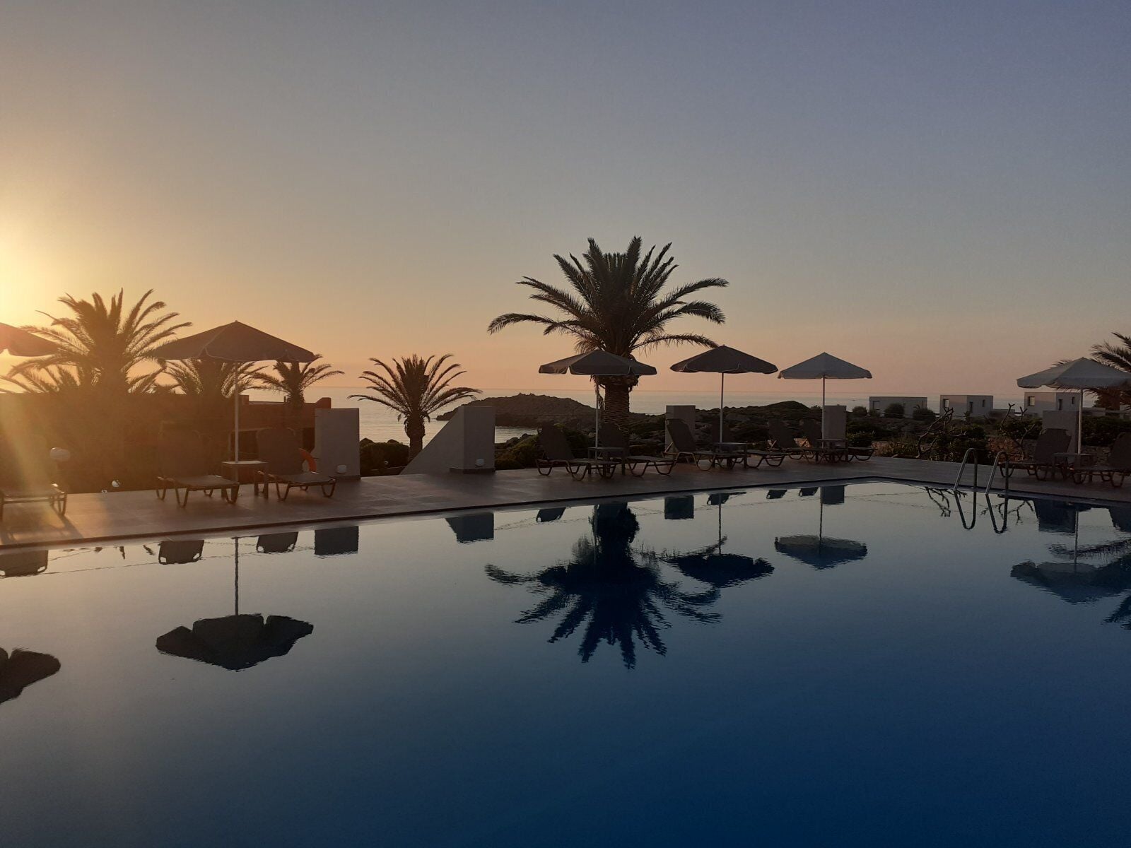 A serene pool area with lounge chairs and umbrellas, surrounded by palm trees and a clear view of the sunset.