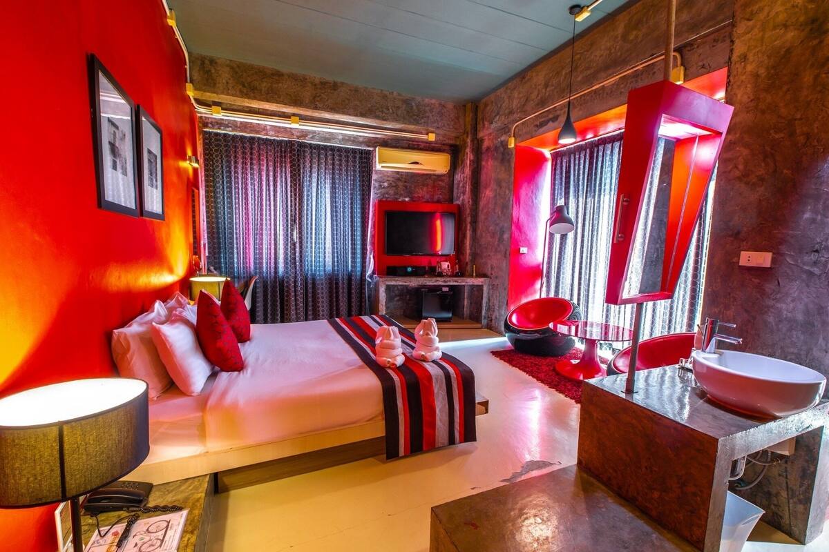 A modern hotel room with a large bed, a TV, a red-framed mirror, and a bathroom with a red sink.