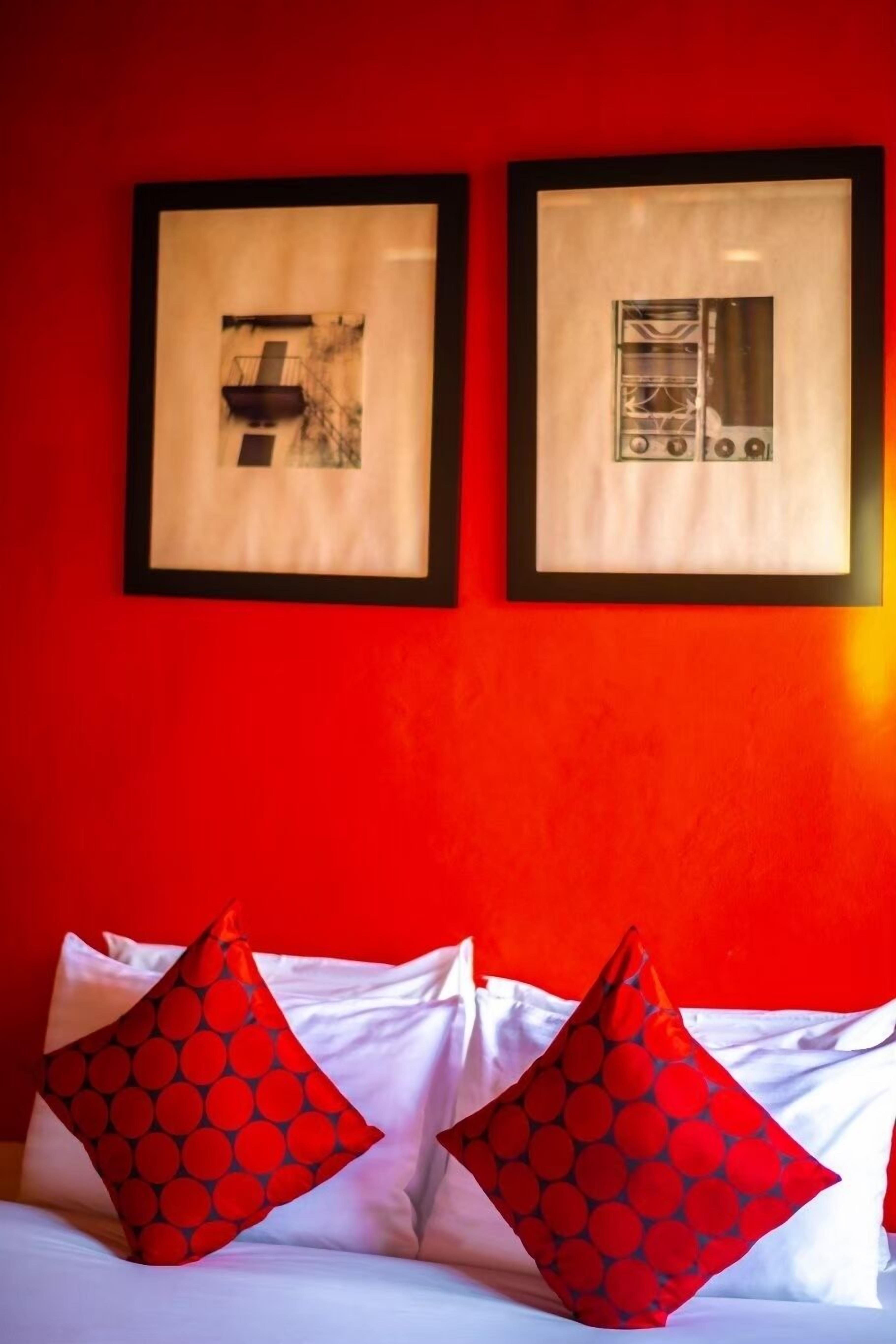A hotel room with a red wall, two framed pictures, and two red pillows with a circular pattern.