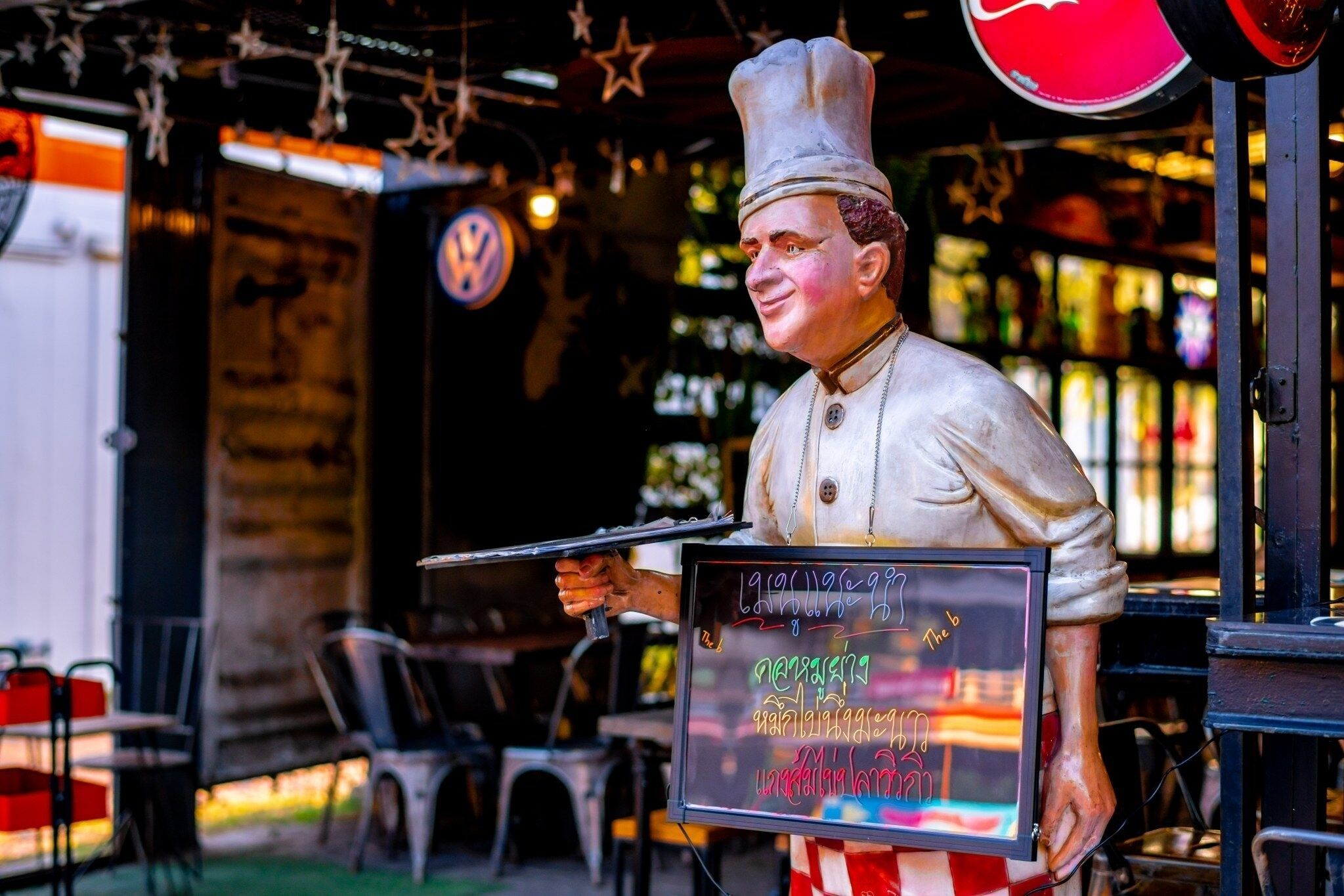 A chef statue holding a chalkboard with menu items, set against a backdrop of outdoor seating and a bar area.