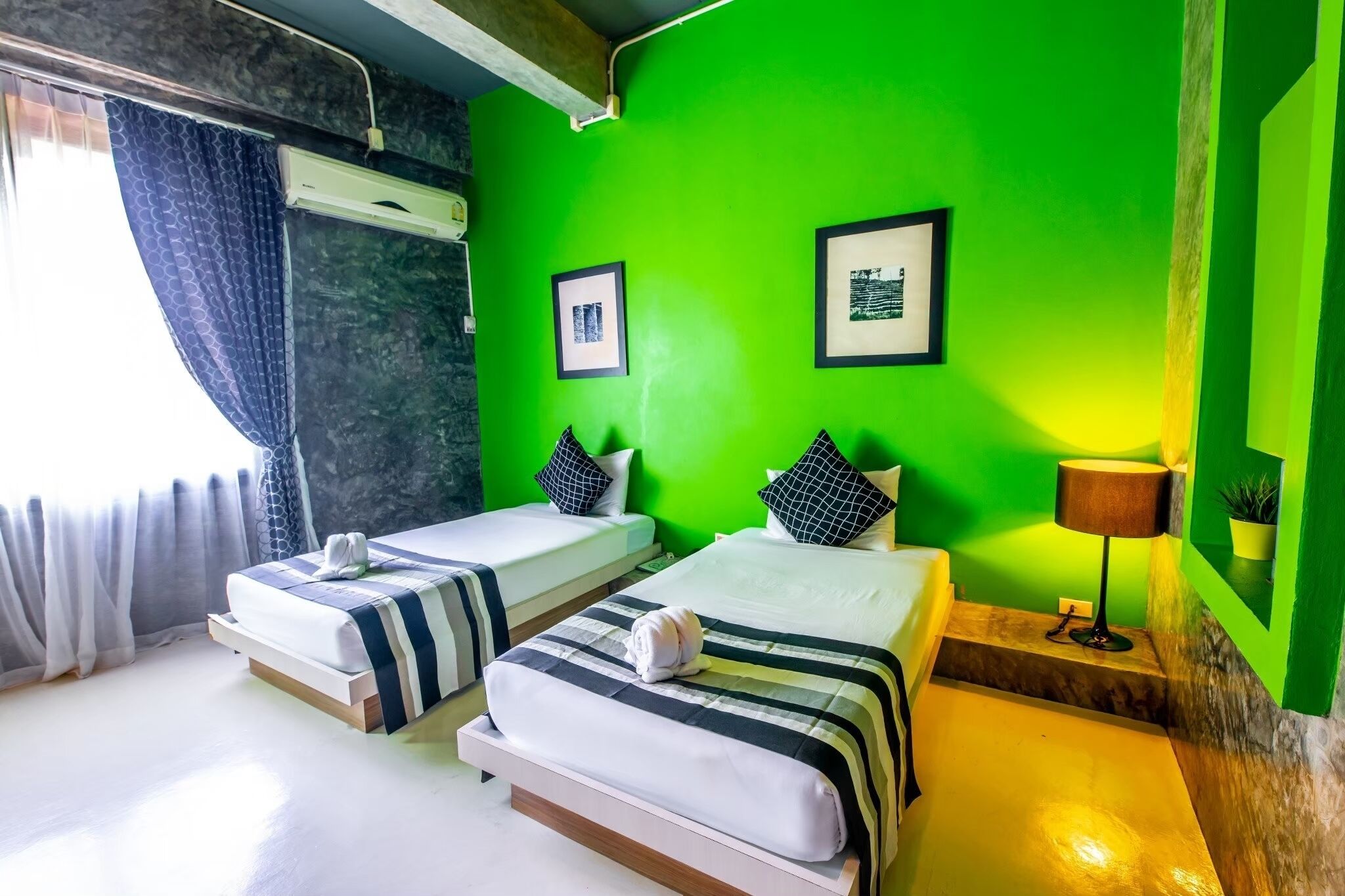 A hotel room with two beds, a green wall, framed pictures, a lamp, and a window with curtains.