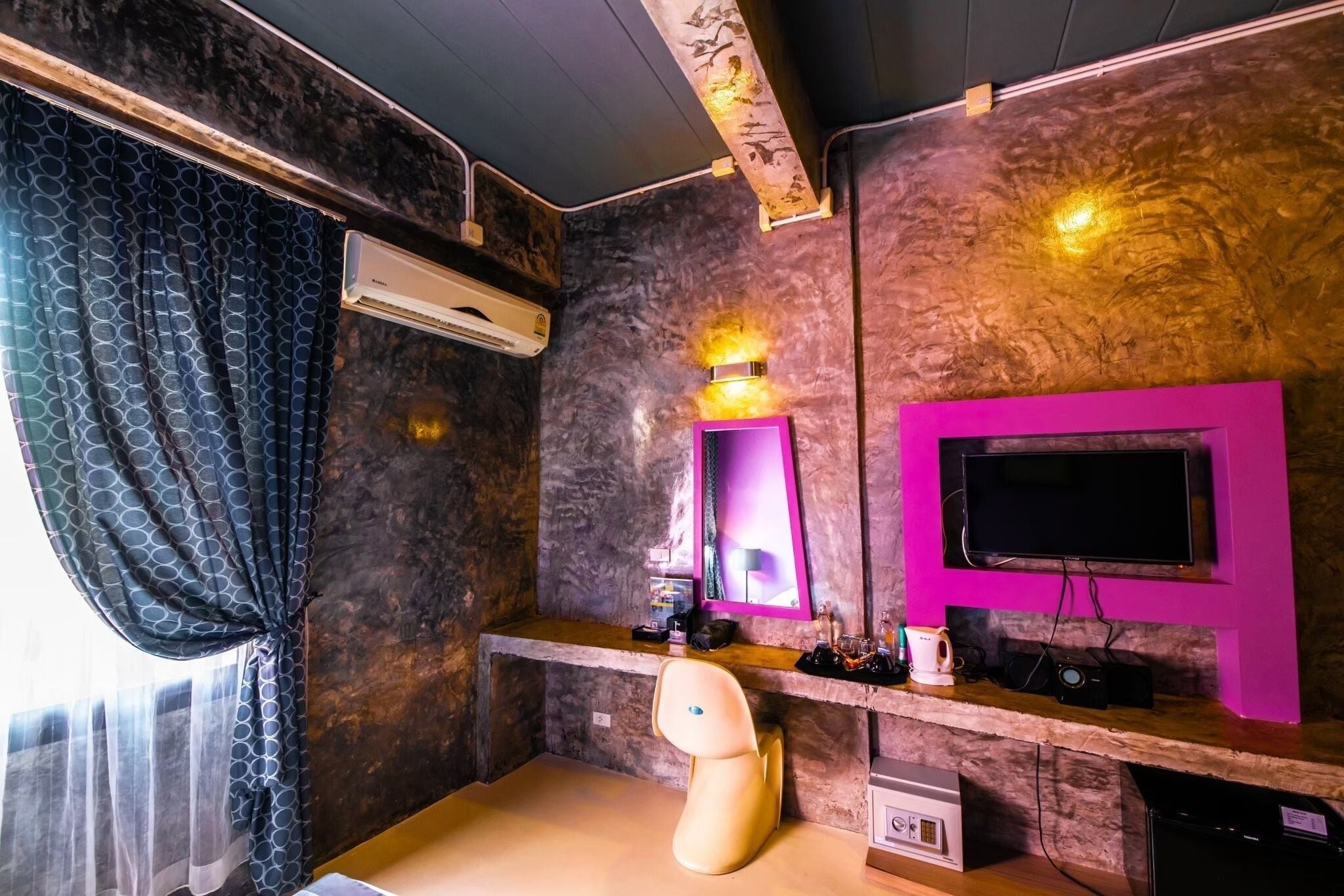 A room with a marble wall, a mounted TV, a desk with a chair, an air conditioner, and a window with patterned curtains.