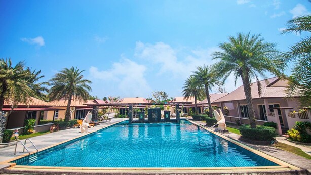 A swimming pool with lounge chairs and palm trees, surrounded by buildings with red roofs. - Pueanjai Resort and Restuarant (Chumphon)