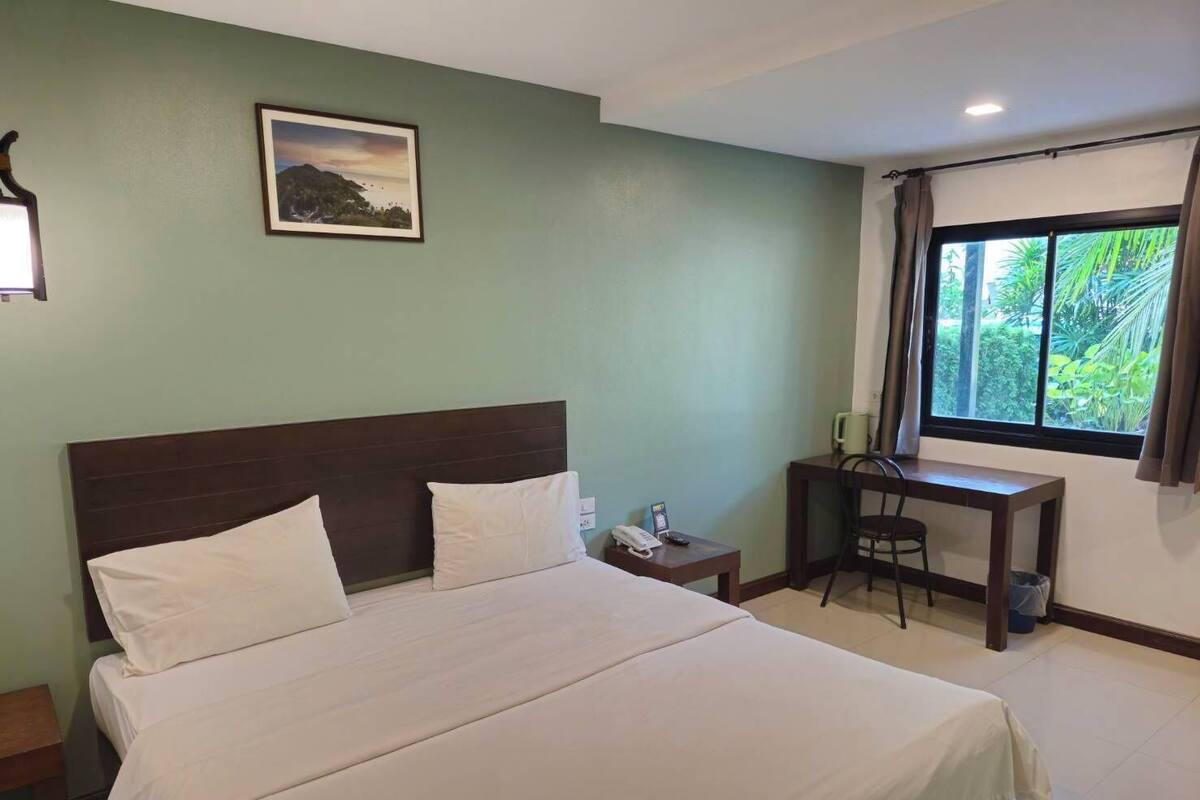 A hotel room with a large bed, a desk, a chair, a nightstand, and a window with a view of greenery.