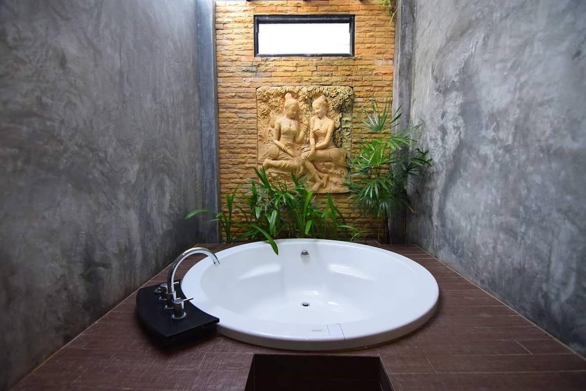 A bathroom with a round white sink, a stone wall with a relief sculpture, and green plants.