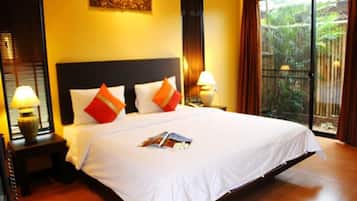 A large bed with white bedding and orange pillows, a nightstand with a lamp, and a view of greenery through a sliding door.
