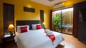 A hotel room with a large bed, two bedside tables with lamps, a window with blinds, a wall-mounted air conditioner, and a view of a bamboo fence and plants.