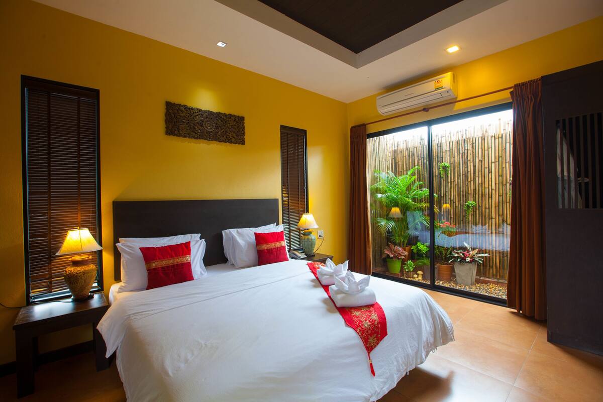A hotel room with a large bed, two bedside tables with lamps, a window with blinds, a wall-mounted air conditioner, and a view of a bamboo fence and plants.