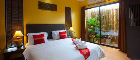 Habitación de hotel con una cama grande, dos mesitas de noche con lámparas, una ventana con persianas, aire acondicionado de pared y vistas a una valla de bambú y plantas.