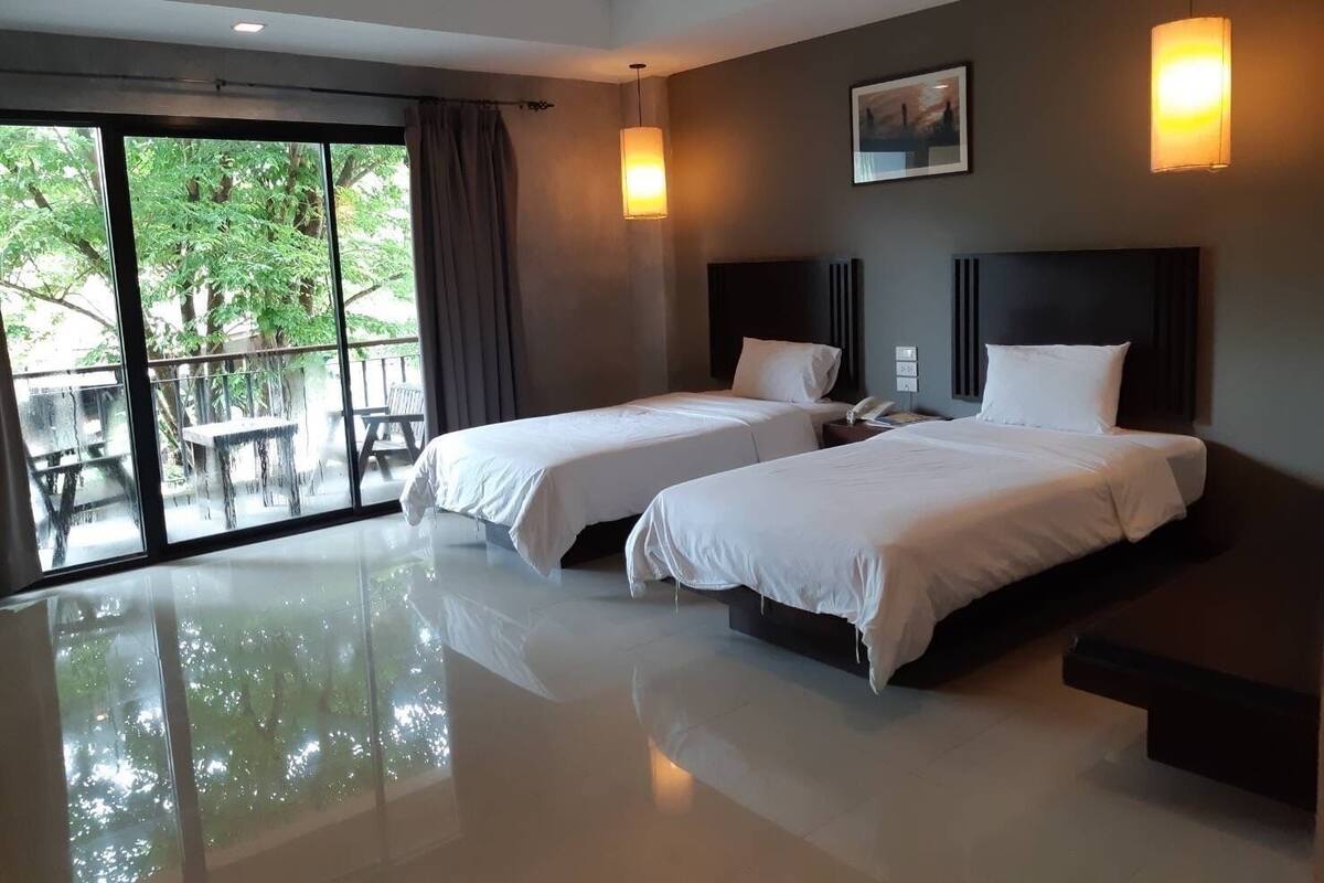 A hotel room with two beds, a large window with a view of greenery, and a balcony.