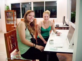 Dos mujeres sentadas en un escritorio blanco, con una computadora portátil, un teclado y un ratón. Se ve una estantería con libros y una planta al fondo.