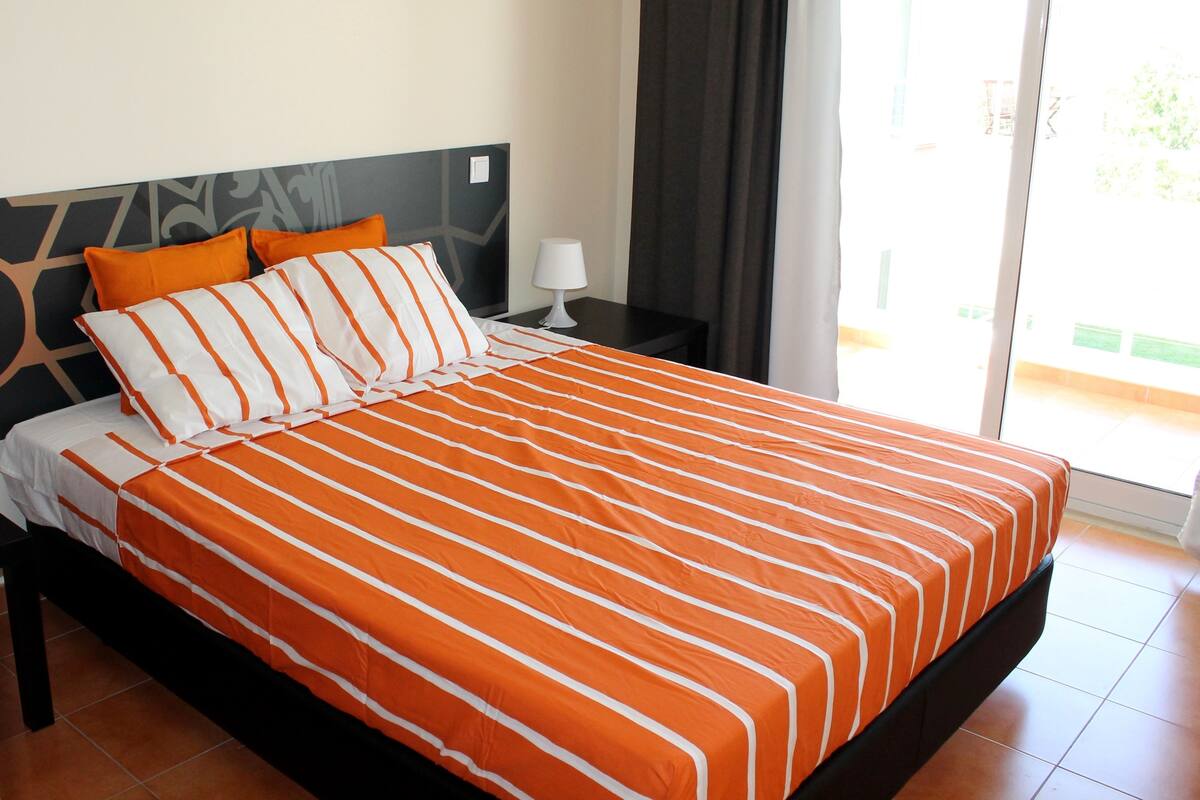 A hotel room with a large bed, orange and white striped bedding, a nightstand with a lamp, and a view of the outdoors through a sliding door.