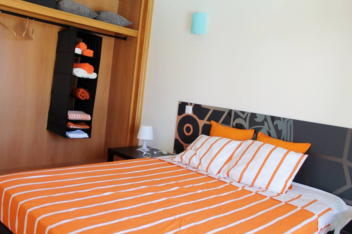 A hotel room with a bed featuring orange and white striped bedding, a headboard with a geometric pattern, and a shelf with folded towels.