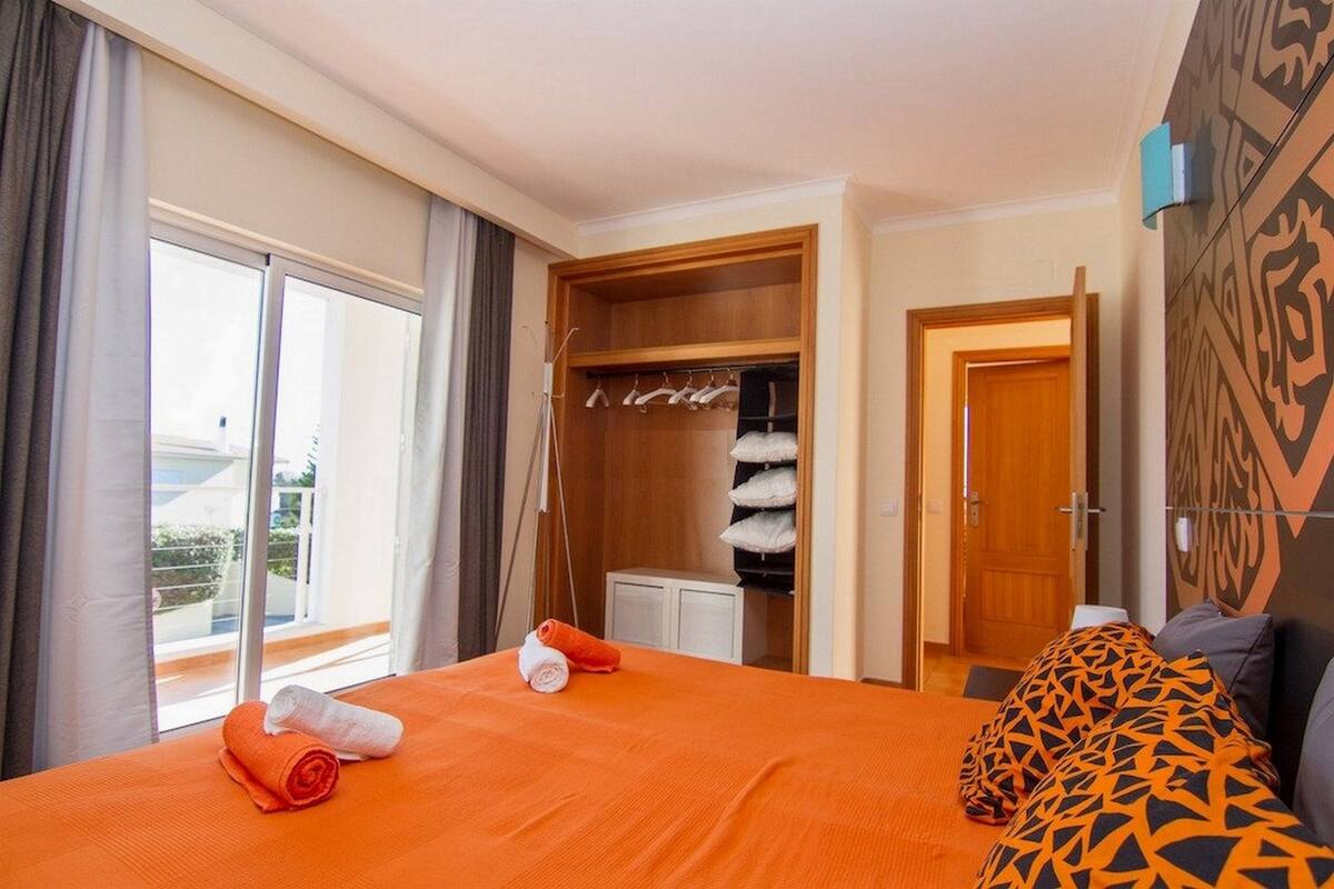A hotel room with a bed, an open closet with hanging clothes, a door leading to another room, and a window with curtains.