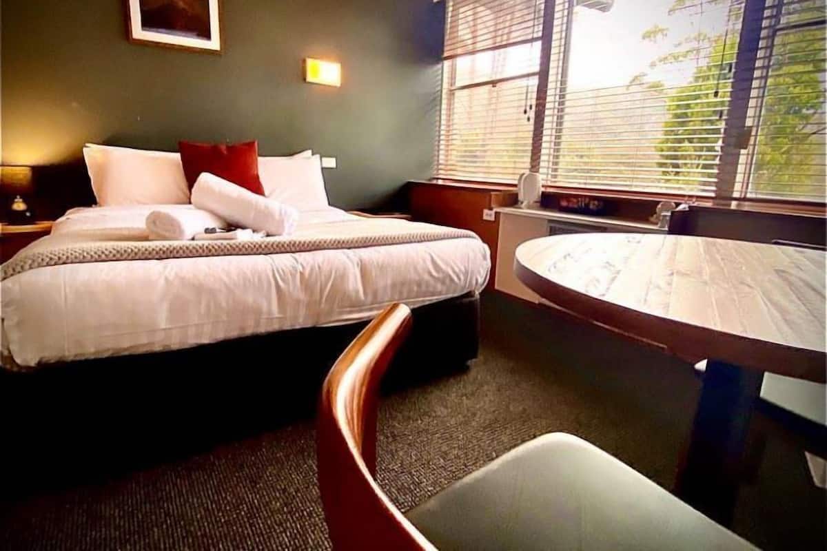 Une chambre d’hôtel avec un lit, une table de chevet, une chaise, une table ronde et une fenêtre avec des stores.