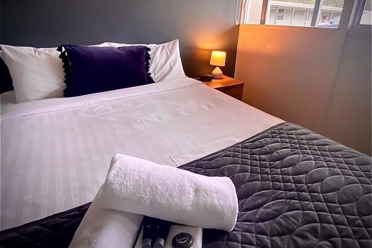 Un lit bien fait, avec des oreillers blancs et foncés, une couverture matelassée et une serviette pliée posée sur le lit. Il y a une table de chevet avec une lampe et une petite bouteille. Un tableau encadré est accroché au mur, au-dessus du lit.