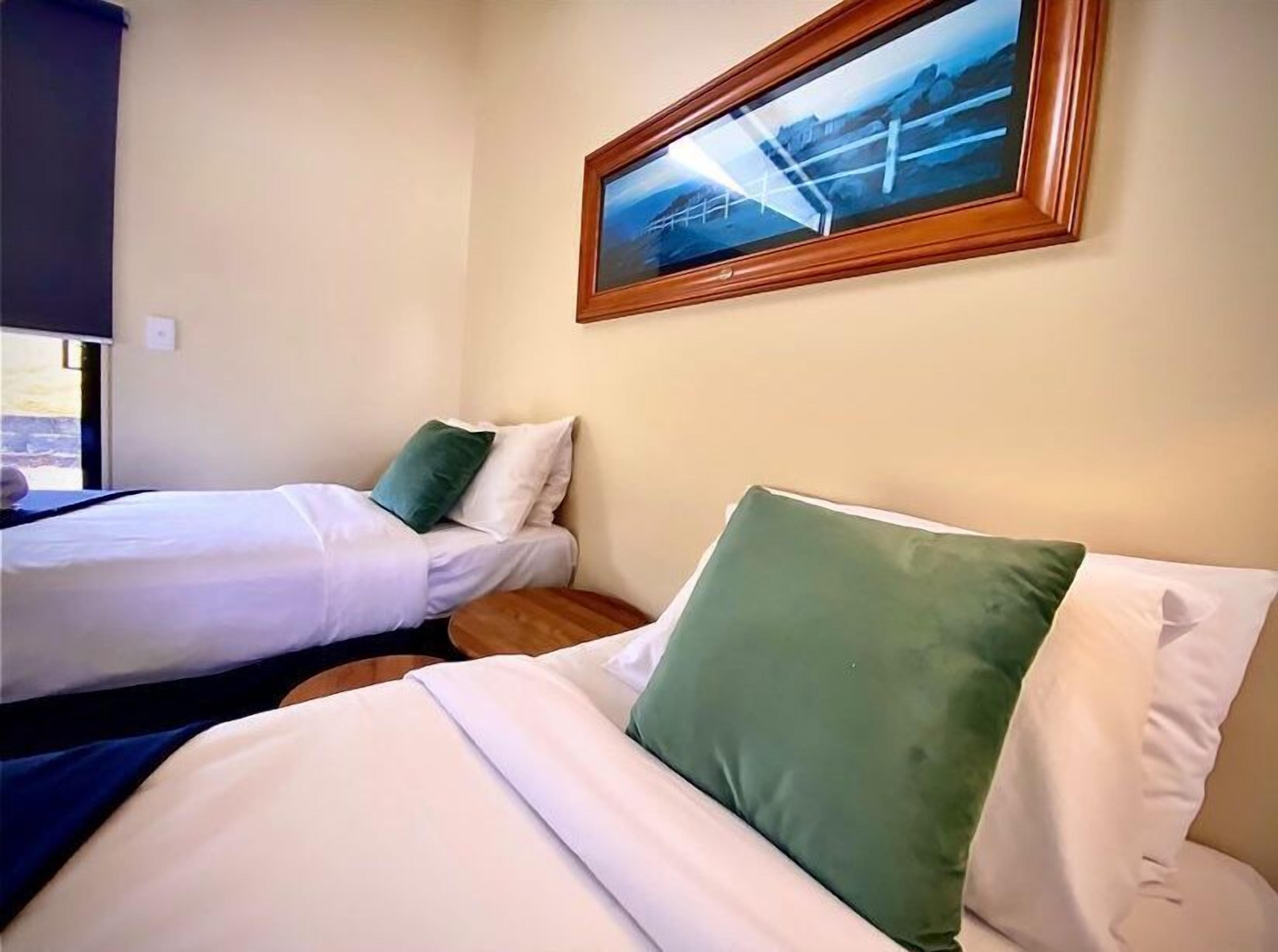 A hotel room with two beds, each with white bedding and green pillows. There is a framed picture on the wall above the beds.