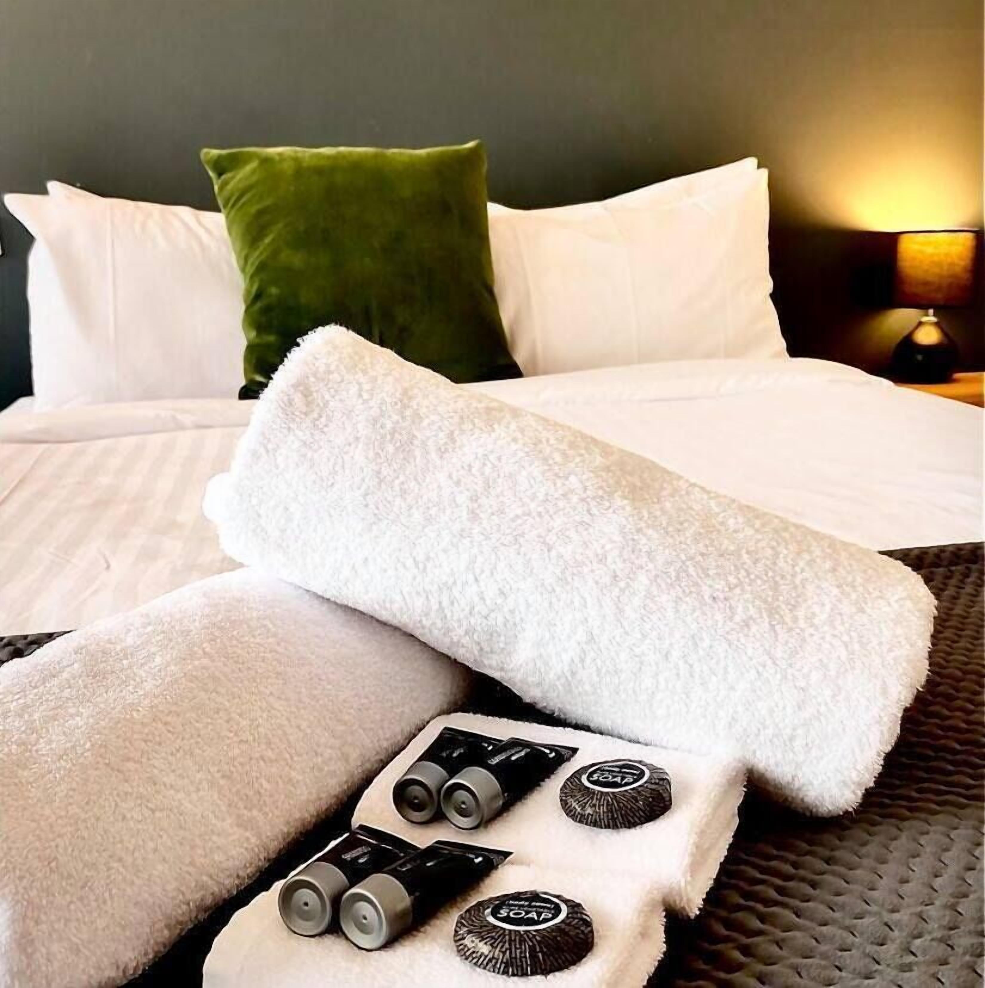A neatly made bed with white linens, a green pillow, and a set of toiletries including soap and lotion.