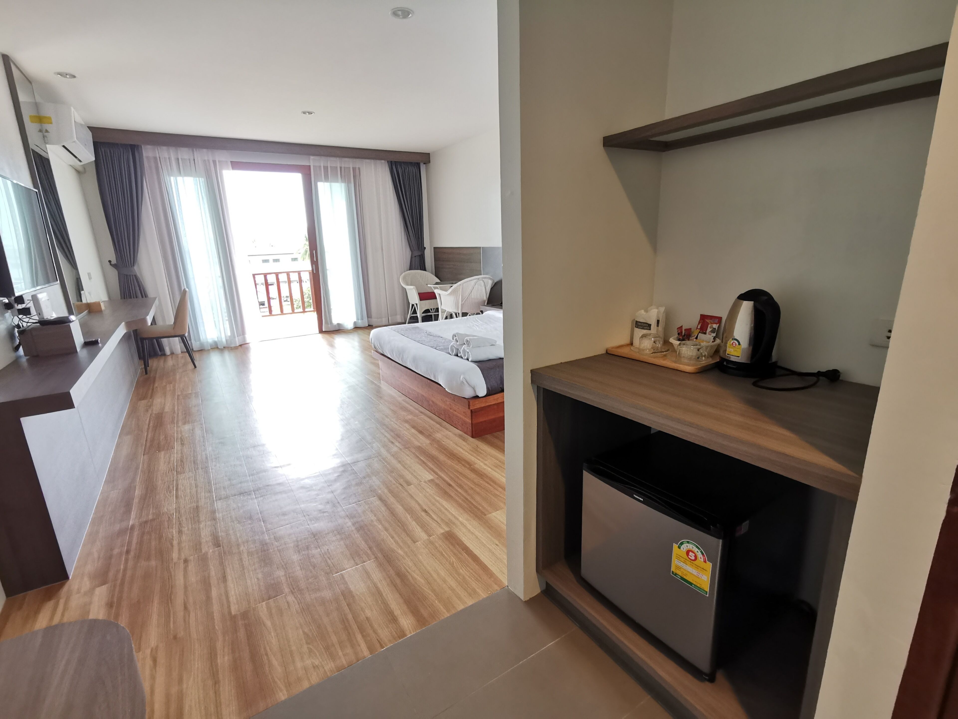A hotel room with a wooden floor, a bed, a desk with a kettle, and a refrigerator.