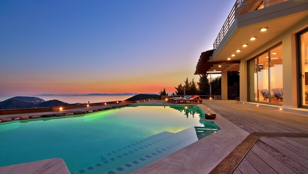 Une piscine avec vue sur les montagnes au crépuscule, une terrasse en bois et un bâtiment moderne doté de grandes fenêtres.