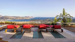 Une terrasse dotée de fauteuils de jardin en osier et de coussins rouges, offrant une vue sur un paysage côtier avec des montagnes et la mer.
