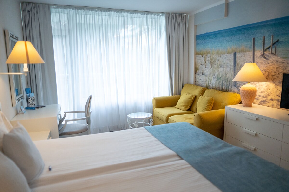 A hotel room with a bed, a yellow sofa, a desk with a lamp, and a beach-themed wall mural.