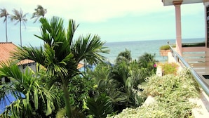 Un balcón con vista al mar, rodeado de plantas tropicales y un edificio con techo de tejas.