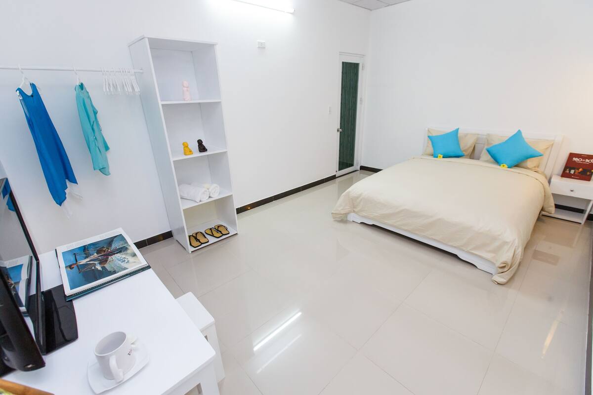 A hotel room with a bed, a white desk with a laptop and a cup, a wardrobe, and a door.