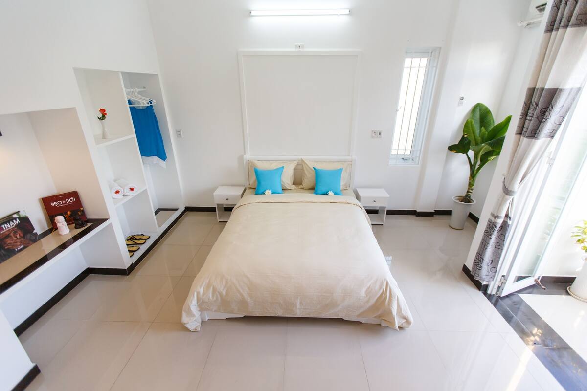 A bedroom with a bed, nightstands, a vanity with a mirror, a shelf with toiletries, a potted plant, and a window with curtains.