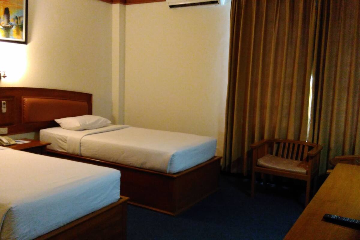 Une chambre d’hôtel avec deux lits simples, une tête de lit en bois, une petite table avec un t éléphone, une chaise et un tableau au mur.