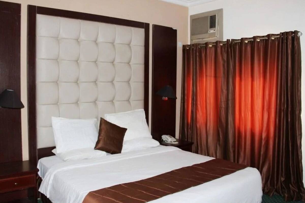 A hotel room with a large bed, a headboard, a bedside table with a lamp, a telephone, and a curtain.