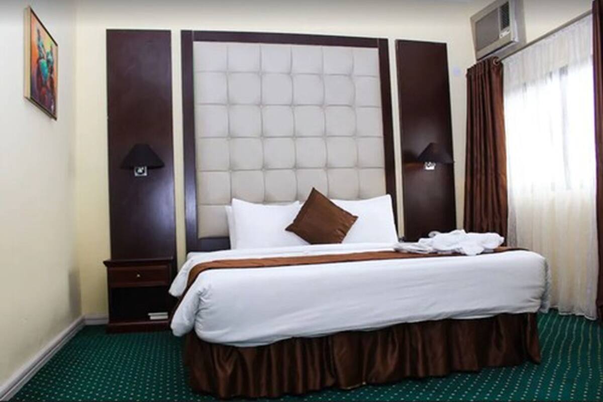 A hotel room with a large bed, two bedside tables, a headboard, and a window with curtains.