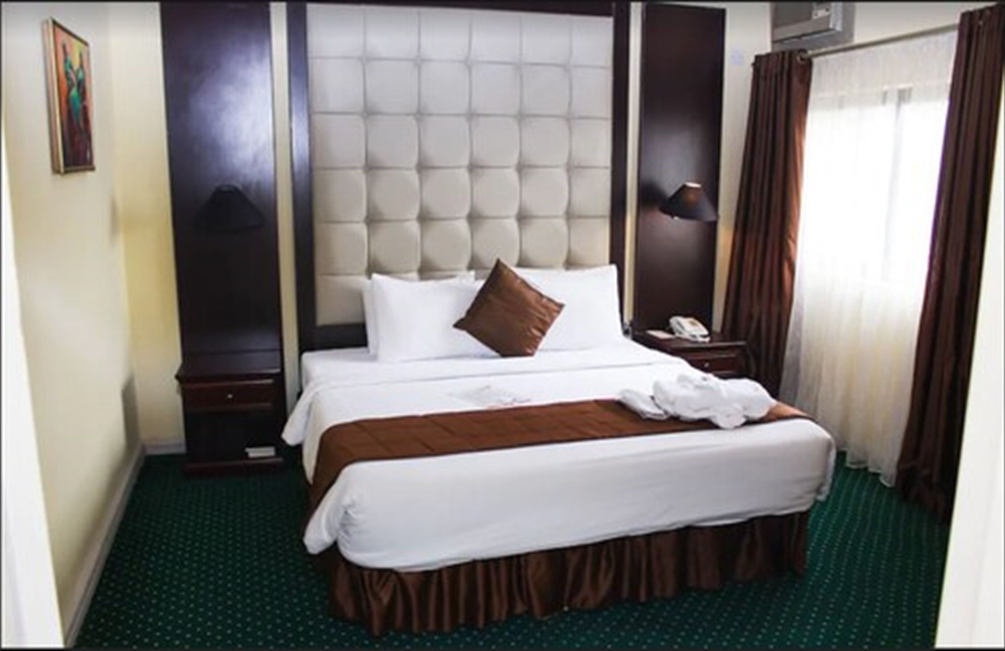 A hotel room with a large bed, a headboard, a bedside table with a lamp, a telephone, and a window with curtains.