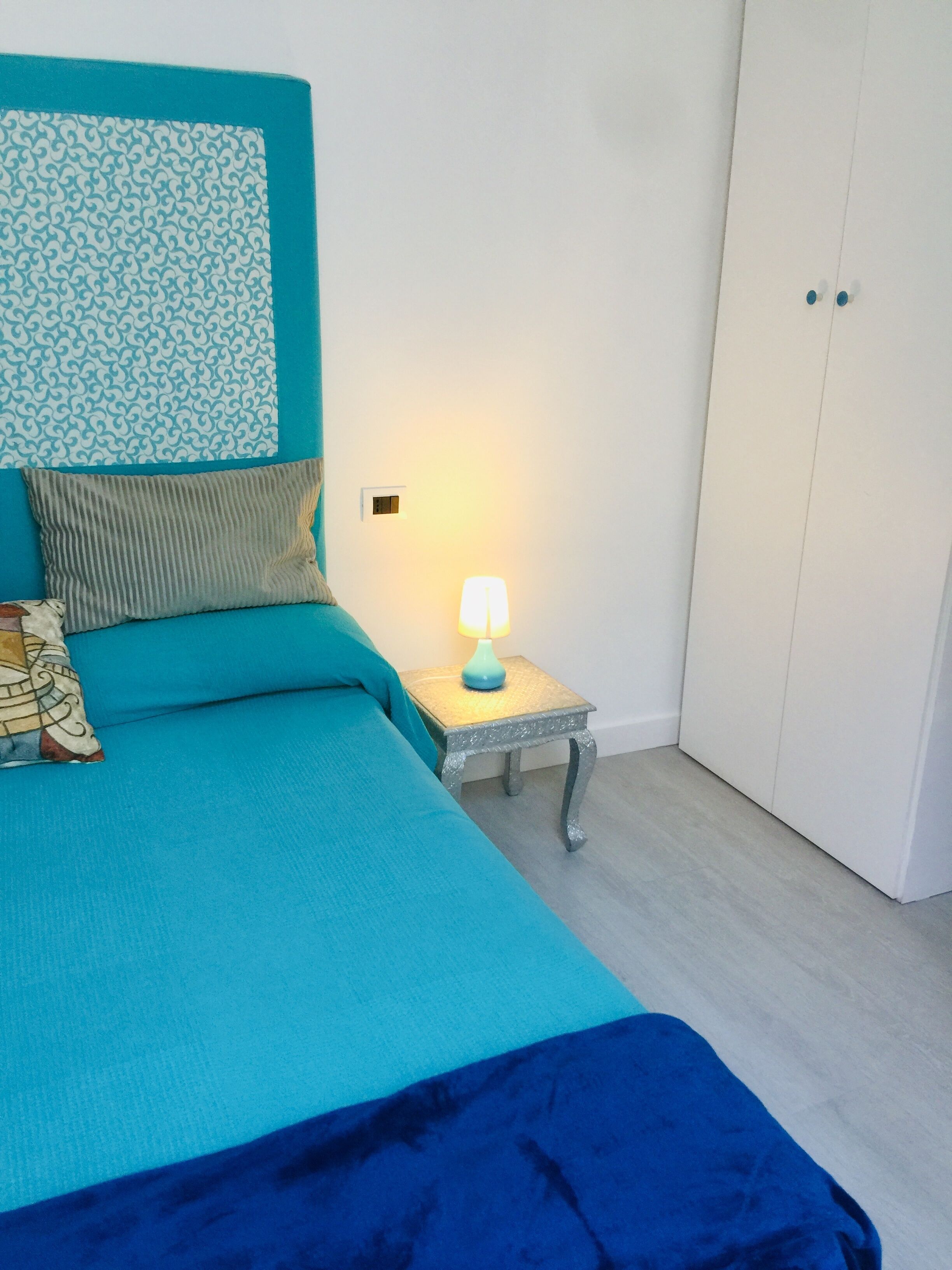 A bedroom with a bed, a nightstand with a lamp, a wardrobe, and a patterned headboard.