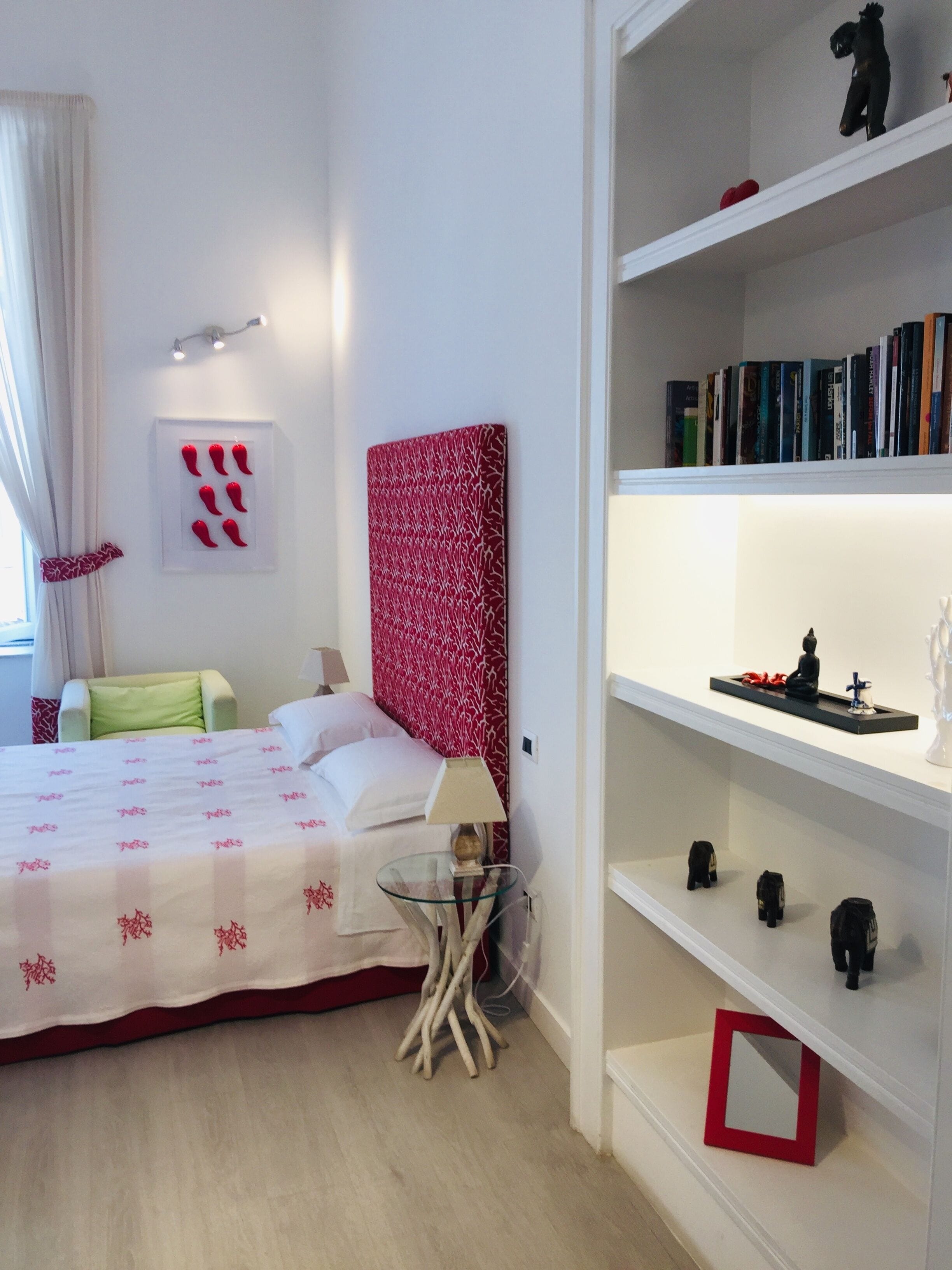A hotel room with a bed, a bedside table with a lamp, a small glass table, a bookshelf with books and decorative items, and a red and white patterned headboard.