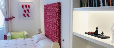 A hotel room with a bed, a bedside table with a lamp, a small glass table, a bookshelf with books and decorative items, and a red and white patterned headboard.