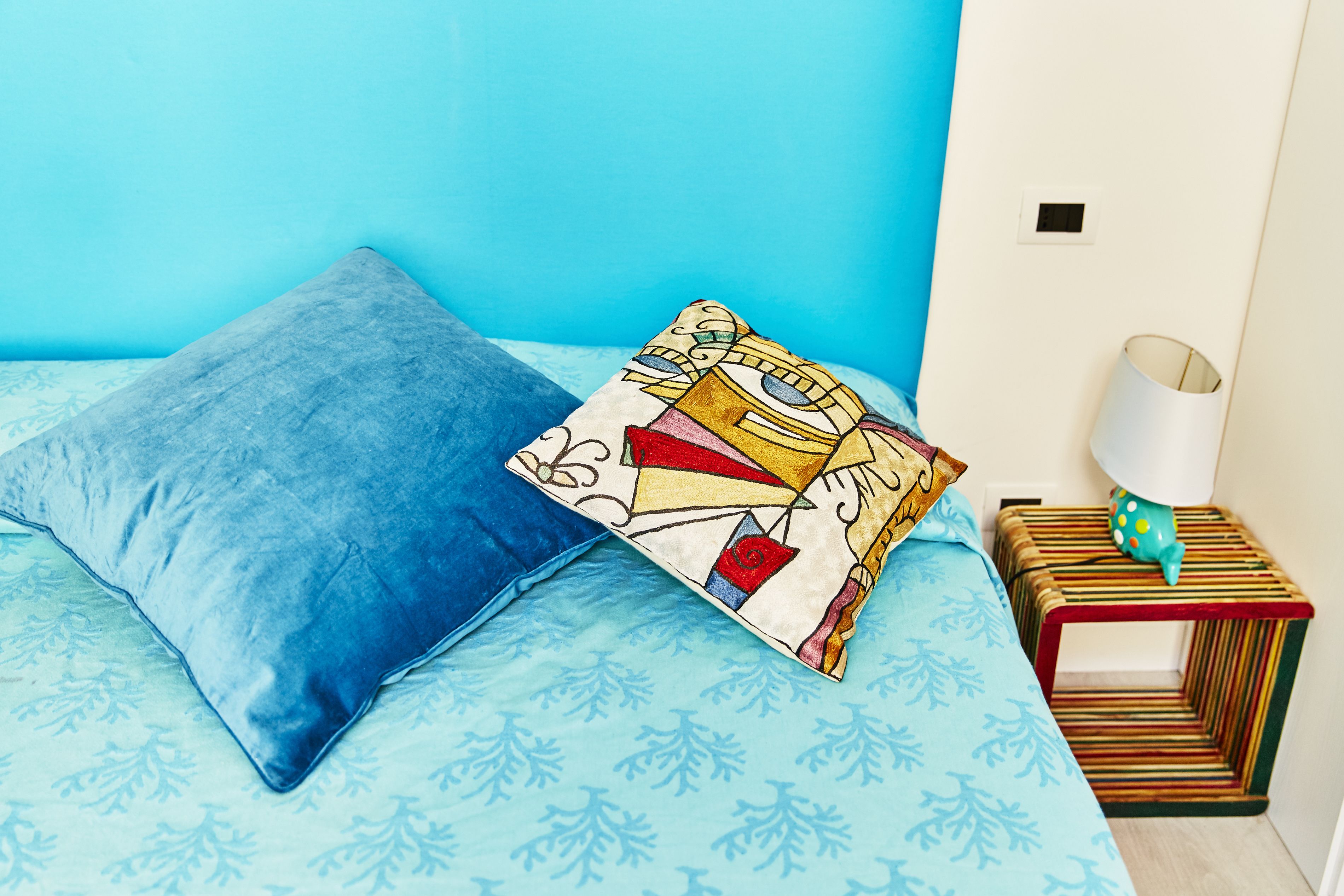 A bed with a blue blanket and pillows, a striped nightstand with a lamp, and a patterned wall.