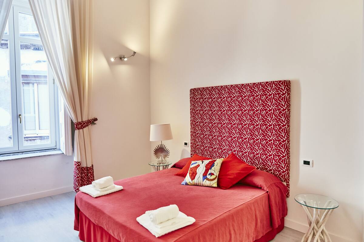 A hotel room with a red bedspread, a patterned headboard, a glass side table with a lamp, and a view of the outdoors through a window with curtains.