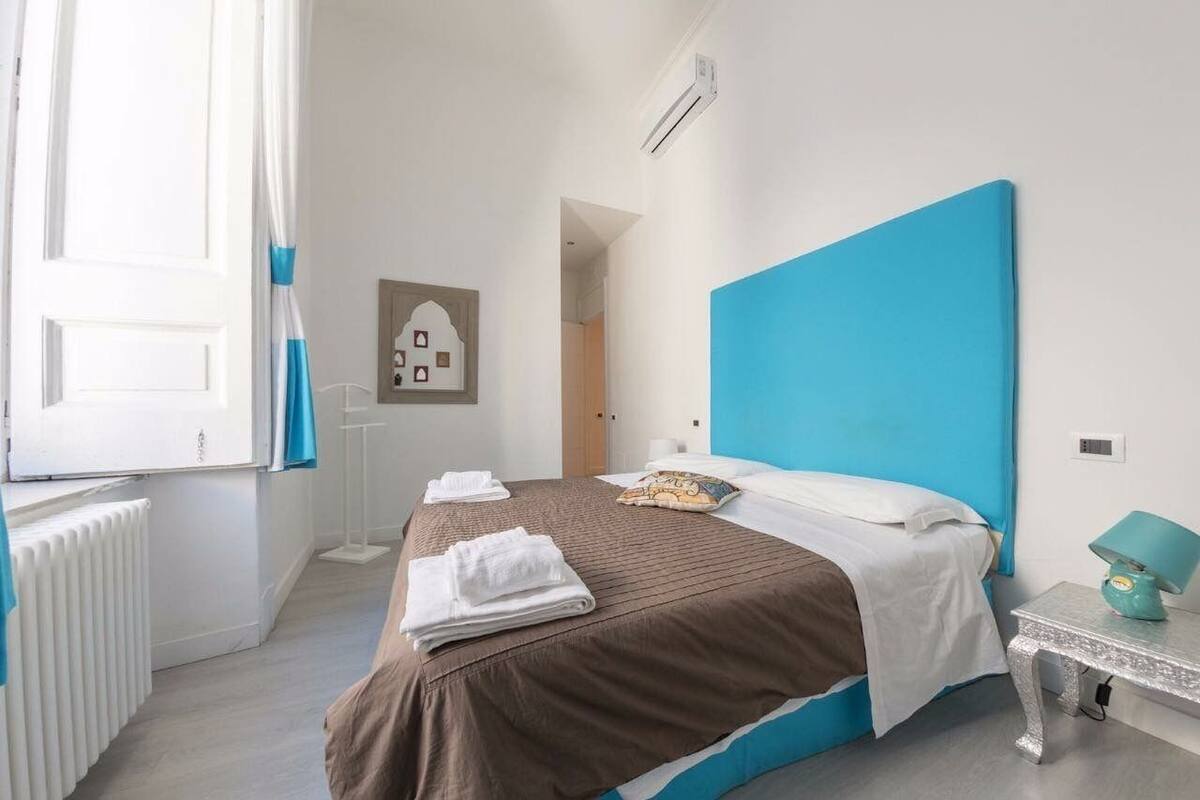A hotel room with a bed, a radiator, a window with curtains, a bedside table with a lamp, and a wall-mounted air conditioner.
