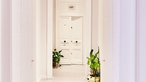 Un pasillo con paredes blancas y piso de madera que conduce a una puerta con un letrero encima. Hay dos plantas en macetas a ambos lados del pasillo.