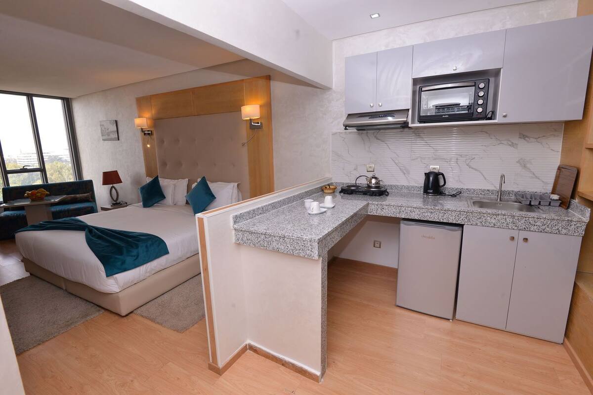 Una habitación de hotel moderna con kitchenette, una cama con cabecera y una zona de estar con sofá y mesa.